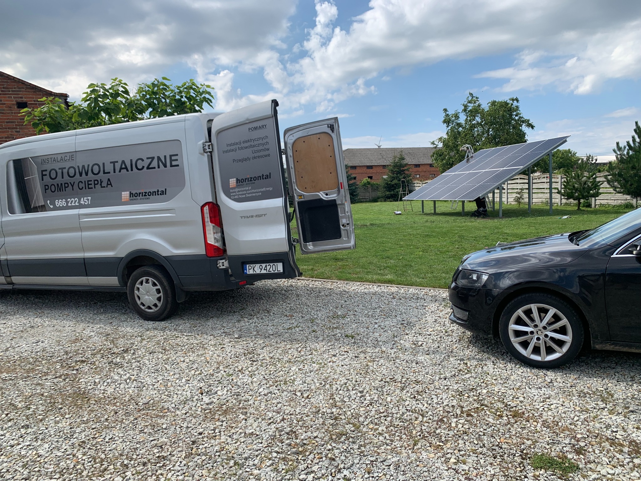 Montaż paneli fotowoltaicznych na trawniku z widocznym samochodem osobowym i busem firmy instalacyjnej z otwartymi drzwiami, w tle budynek gospodarczy.