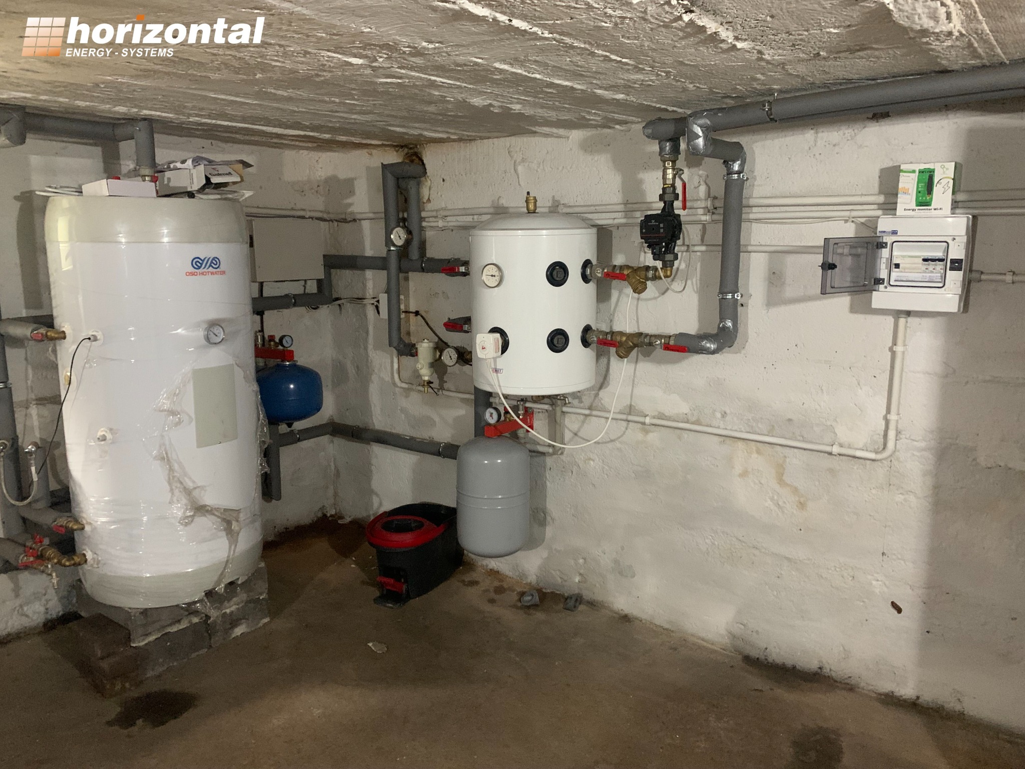 Dwa białe zbiorniki ciepłej wody użytkowej w piwnicy, z widocznymi rurami, manometrami i zaworami, oraz skrzynka elektryczna z licznikiem energii na ścianie.