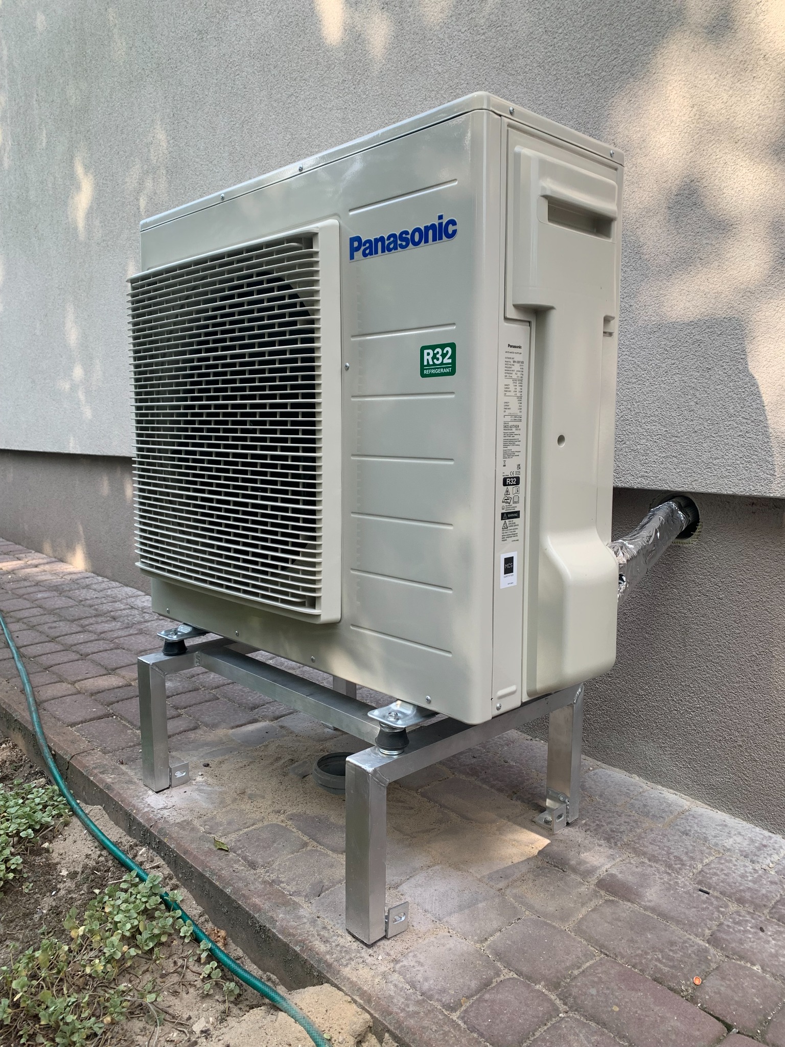 Zewnętrzna jednostka klimatyzacji Panasonic R32 na metalowej podstawie, zamontowana przy szarej ścianie budynku w Nowych Skalmierzycach. Widoczny przewód odprowadzający skropliny.