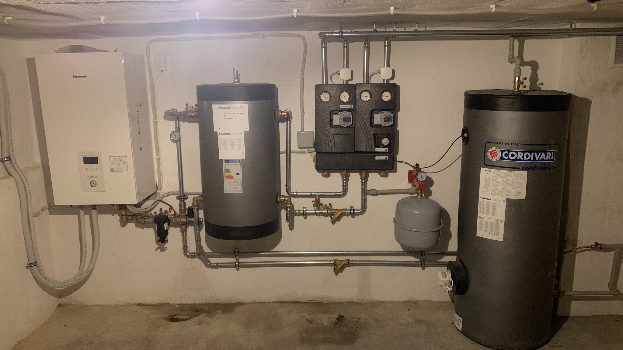 Kompletny system grzewczy z białą jednostką Panasonic Aquarea, dwoma szarymi zbiornikami Cordivari i rozbudowaną instalacją rur w pomieszczeniu technicznym.