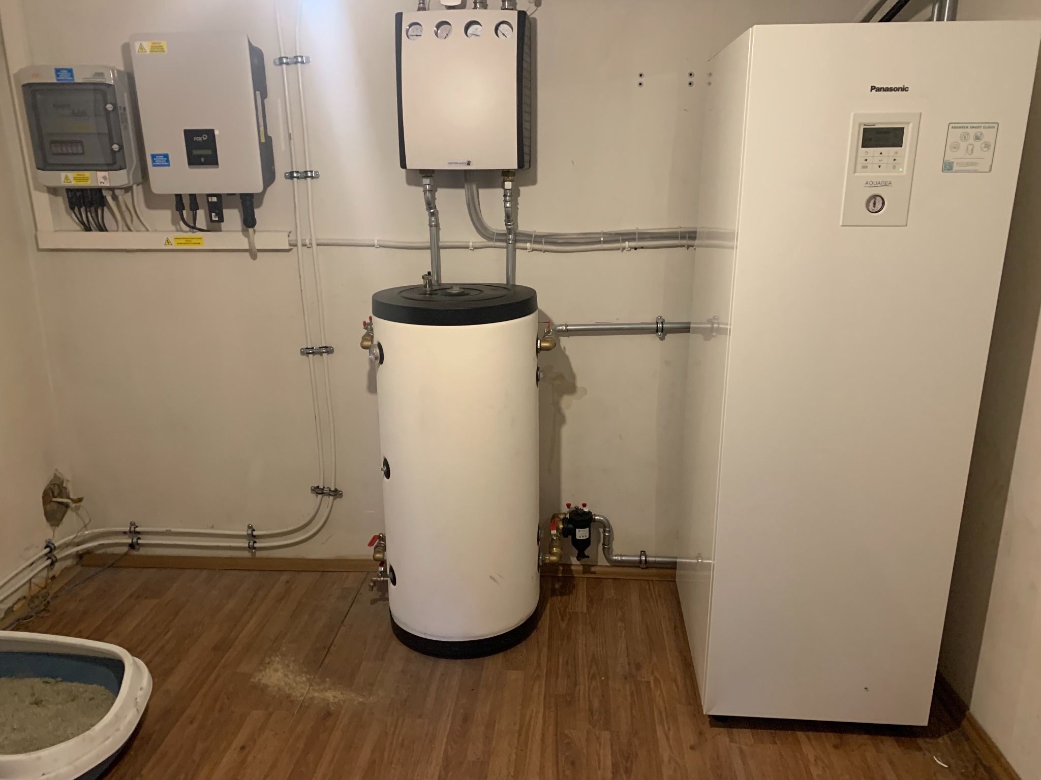 Biała pompa ciepła Panasonic Aquarea, zasobnik ciepłej wody, falownik fotowoltaiczny i licznik energii elektrycznej zamontowane w pomieszczeniu z drewnopodobną podłogą.