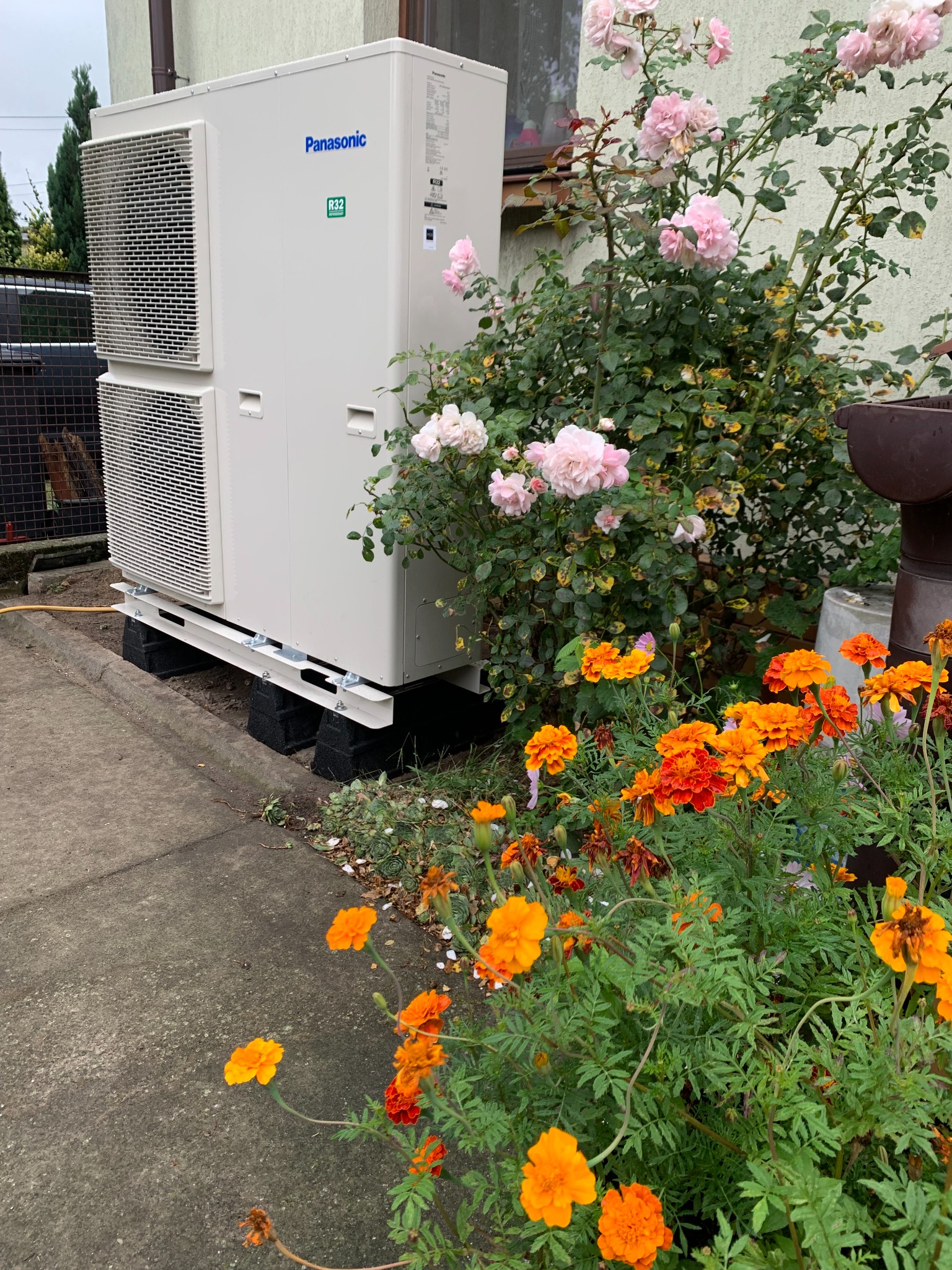 Zewnętrzna jednostka dwuczęściowej pompy ciepła Panasonic, zainstalowana na betonowych blokach, otoczona kwitnącymi nagietkami i różowymi różami.