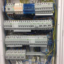 Wnętrze skrzynki elektrycznej z zainstalowanymi bezpiecznikami, przewodami w kolorach żółtym, niebieskim i czarnym, oraz elementami zabezpieczeń przeciwprzepięciowych.