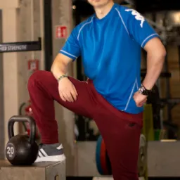 Mężczyzna w sportowym stroju, opierający nogę na skrzyni z odważnikiem 20 kg w siłowni, z widocznym sprzętem do ćwiczeń w tle.