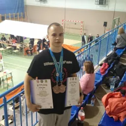 Młody mężczyzna z medalami i dyplomami na tle hali sportowej podczas zawodów w podnoszeniu ciężarów.