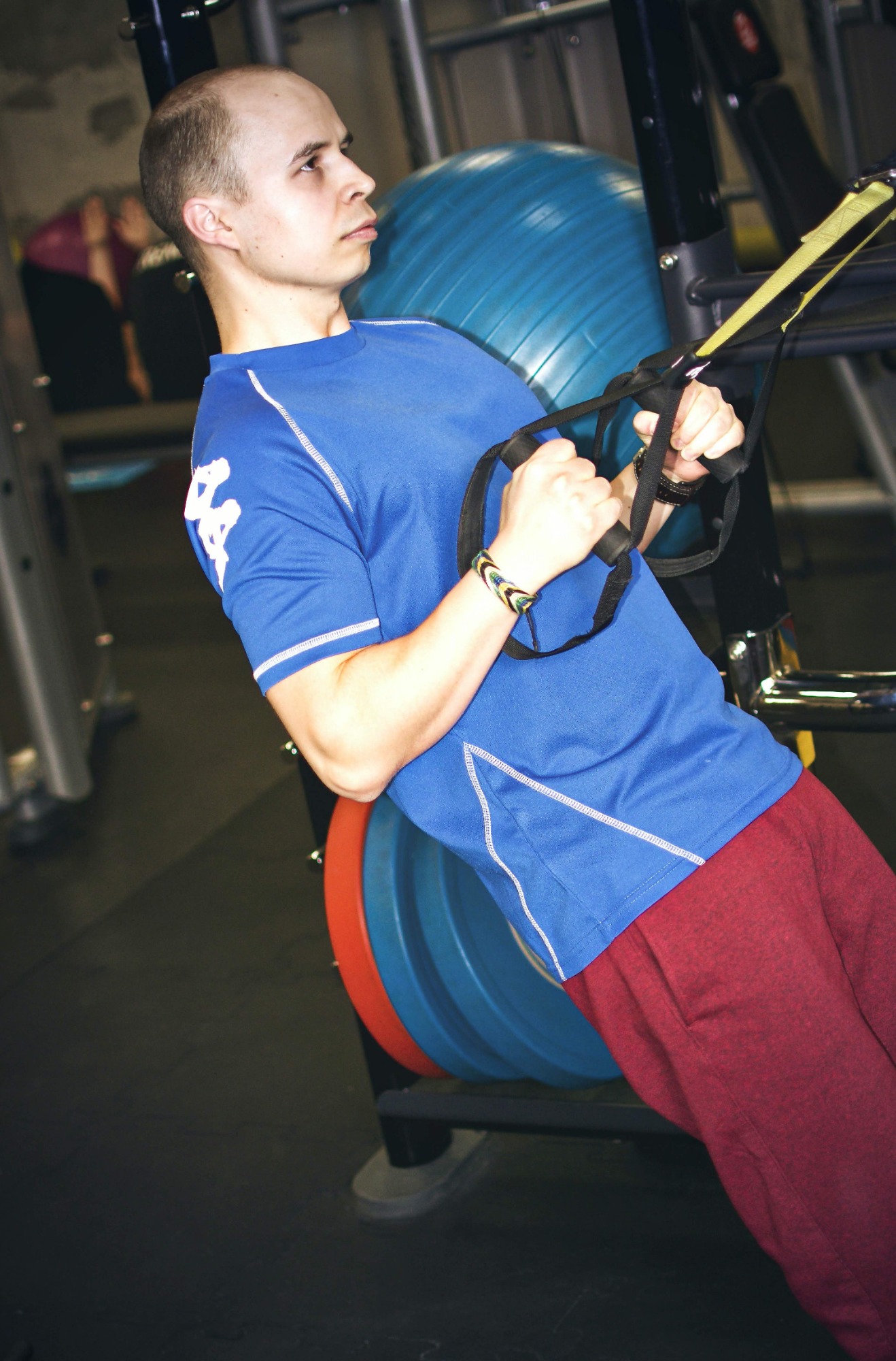 Mężczyzna w niebieskiej koszulce i bordowych spodniach ćwiczy na siłowni, opierając się o piłkę gimnastyczną i używając systemu zawieszeń TRX.