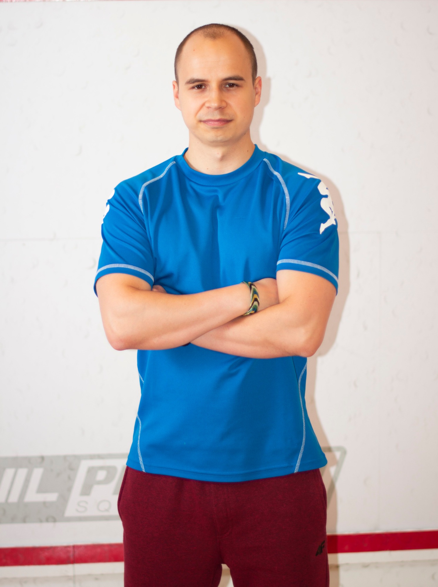 Mężczyzna w sportowej koszulce i spodniach dresowych, z założonymi rękami, stojący na tle ściany kortu do squasha.