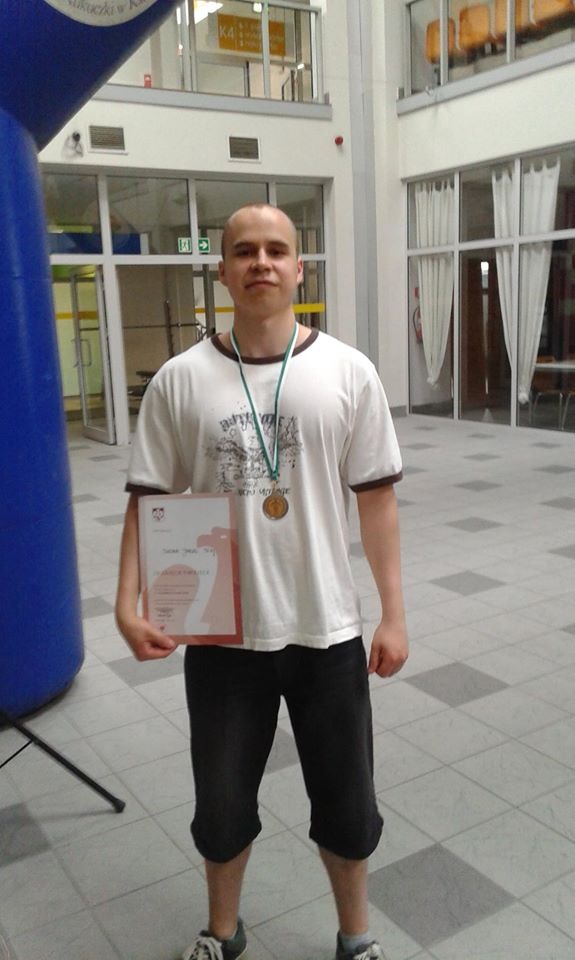 Młody mężczyzna z medalem na szyi i dyplomem w dłoni, stojący w hali sportowej