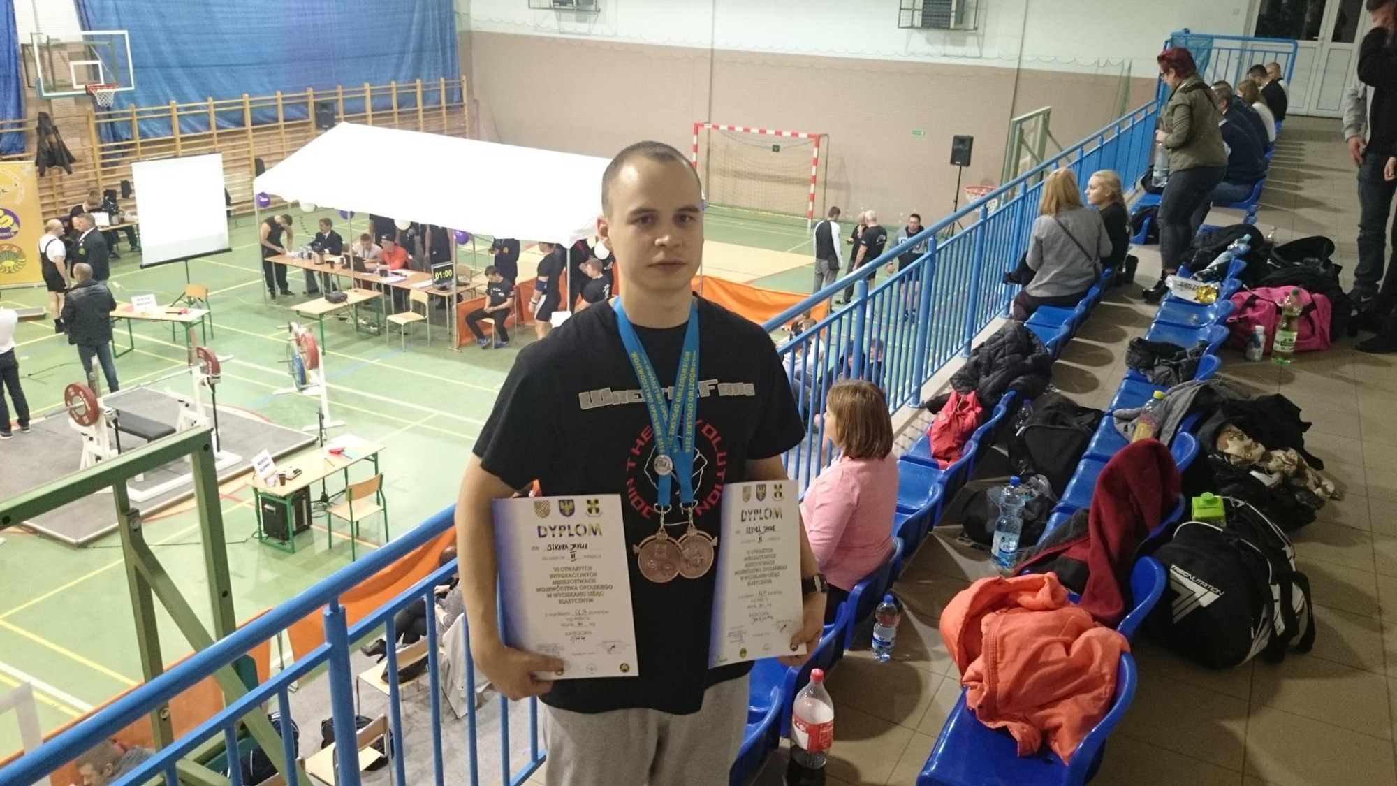 Młody mężczyzna z medalami i dyplomami na tle hali sportowej podczas zawodów w podnoszeniu ciężarów.