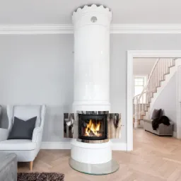 Biały, cylindryczny piec kaflowy z otwartymi drzwiczkami, płomieniami wewnątrz paleniska i dekoracyjną koroną na górze, umieszczony w salonie z parkietem w jodełkę i widocznymi schodami.