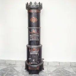 Stary piec żeliwny z dekoracyjnymi elementami z miedzi, zwieńczony koroną, stoi na tle białej ściany i szarej podłogi z płytek.