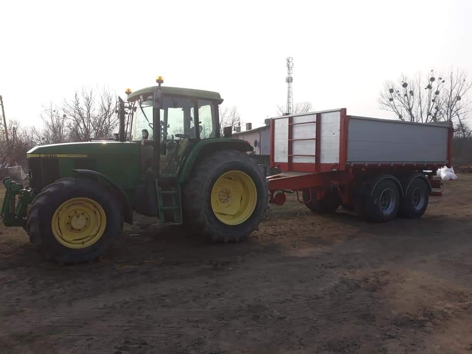 Zielony traktor John Deere z żółtymi felgami, podłączony do szarej przyczepy z czerwonymi bokami, stoi na ziemi w pochmurny dzień.
