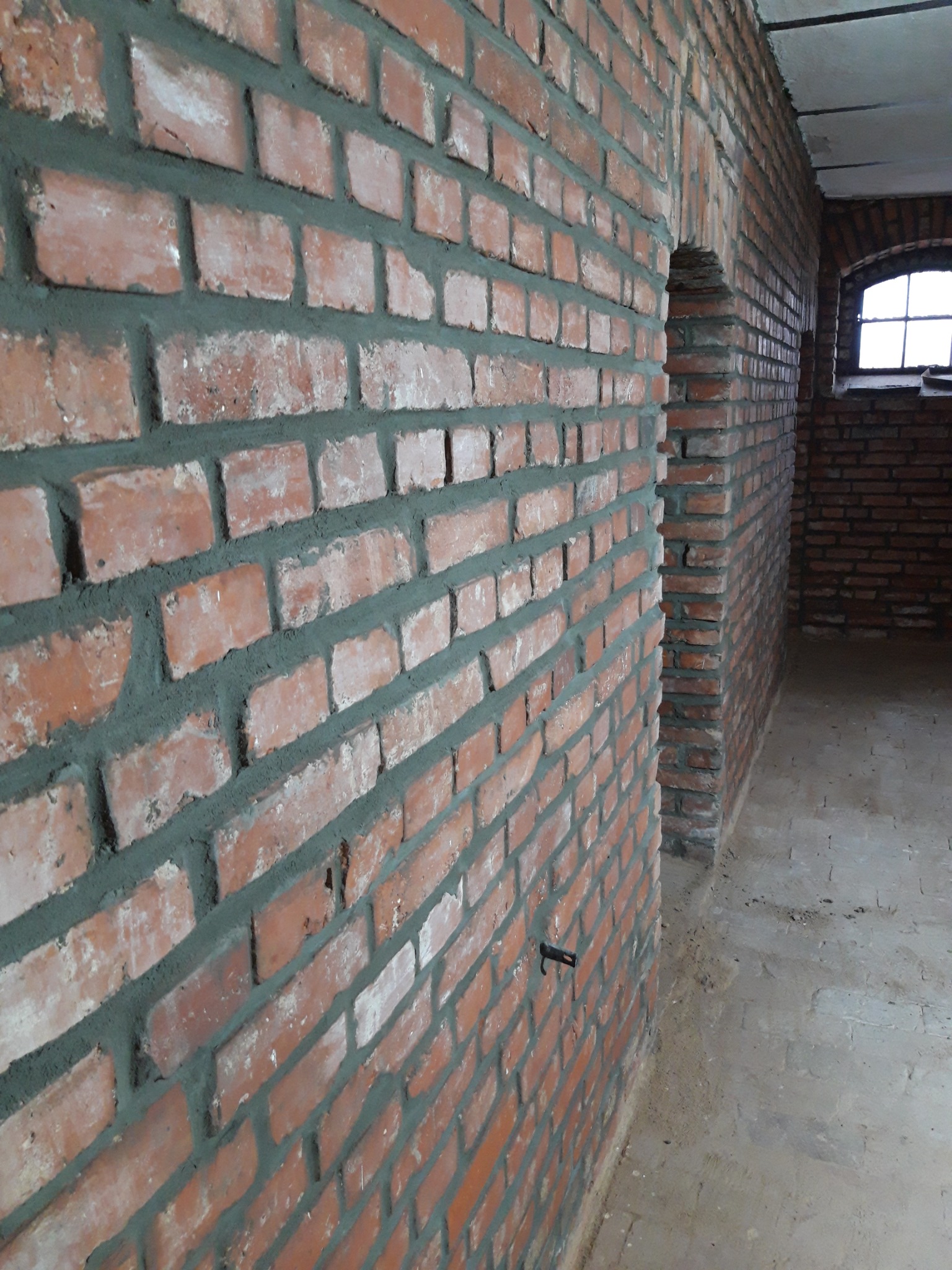 Surowa ściana z czerwonej cegły z szarą fugą, widoczny fragment sklepienia łukowego i okno w głębi pomieszczenia.