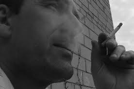 Czarno-białe zdjęcie mężczyzny z papierosem w dłoni, na tle ceglanej ściany. Widoczny fragment twarzy i dłoni, z dymem unoszącym się z papierosa.