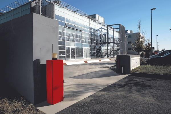 Nowoczesny budynek z przeszkloną fasadą i czerwoną obudową sterownika bramy wjazdowej, widoczne anteny i lampka sygnalizacyjna.