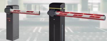 Dwie automatyczne szlabany parkingowe z ramionami w kolorze biało-czerwonym, widoczne na tle rozmytego otoczenia budynku.