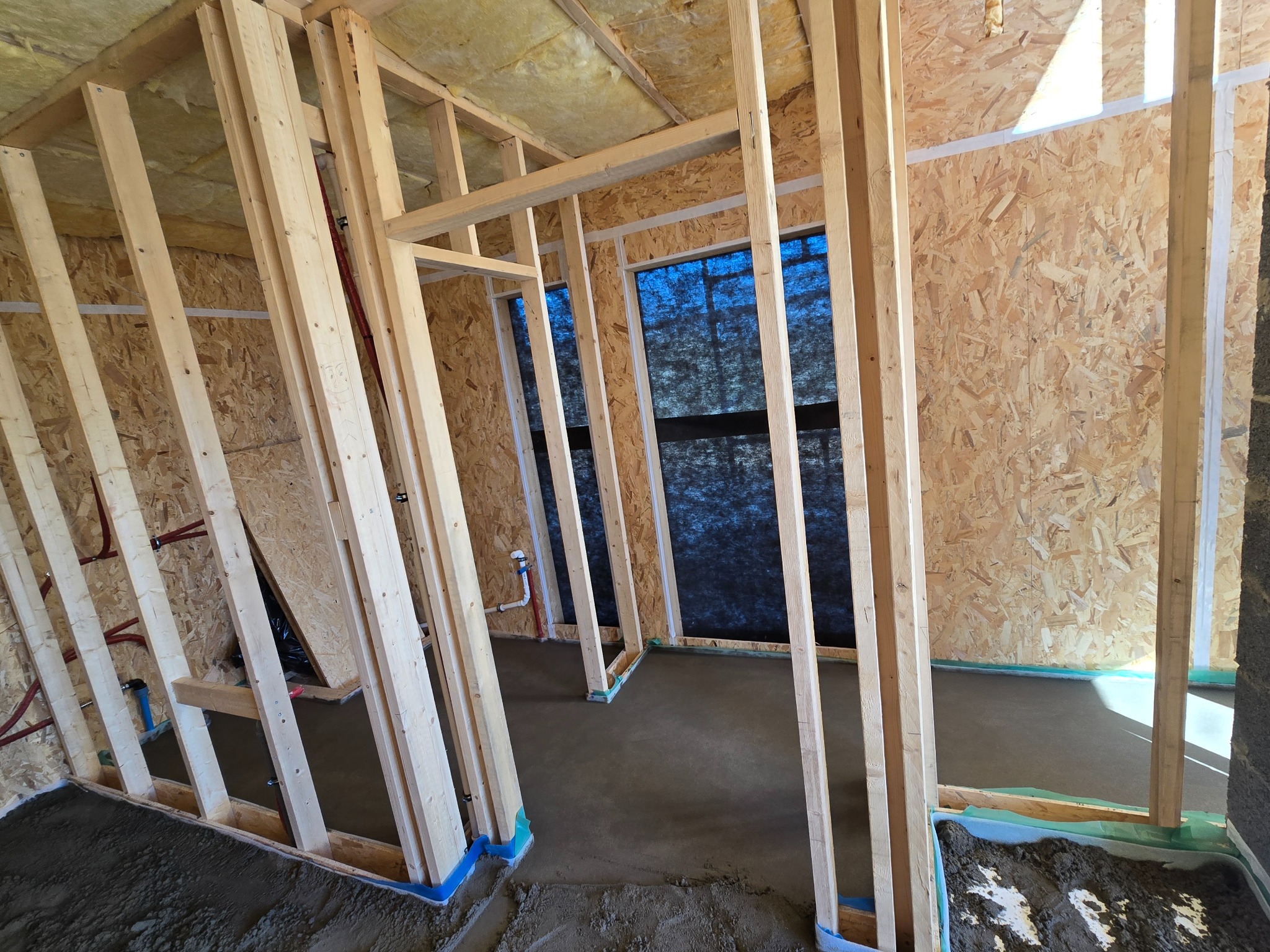 Wnętrze w trakcie budowy z drewnianą konstrukcją ścian i wylaną, szarą posadzką jastrychową, zabezpieczoną folią. Widoczne instalacje i ocieplenie stropu.