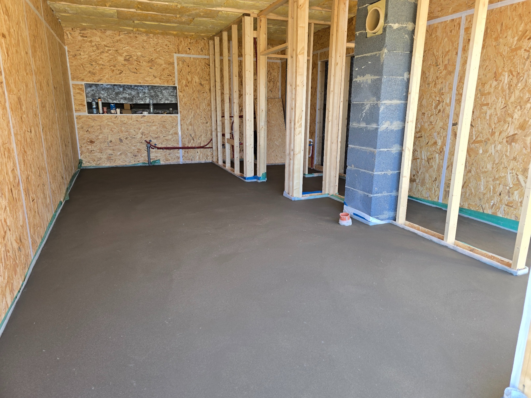 Wylana, gładka posadzka jastrychowa w surowym wnętrzu z drewnianą konstrukcją ścian i oknem. Widoczny komin z szarych pustaków.