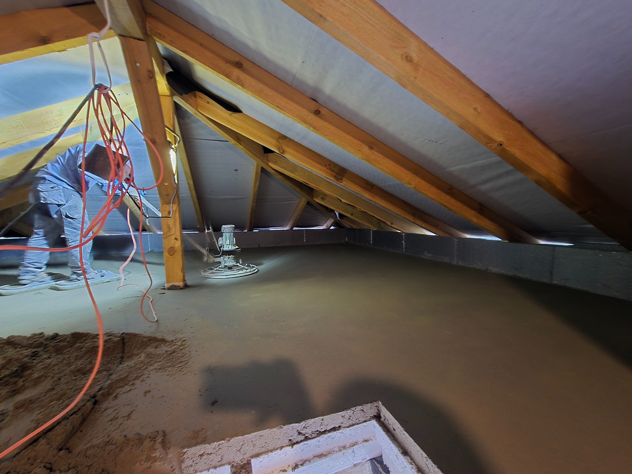 Praca przy wylewaniu posadzki jastrychowej na poddaszu, widoczna maszyna do zacierania i fragment osoby w roboczym ubraniu. Elementy konstrukcji dachu w tle.