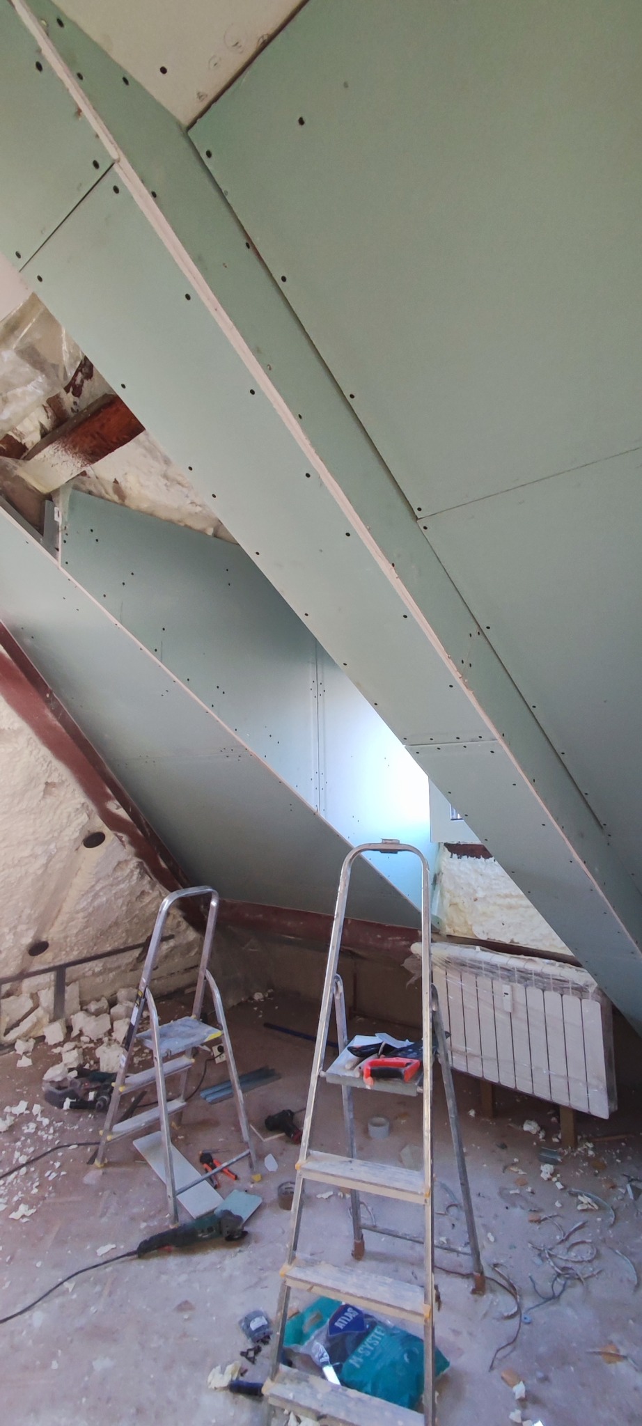 Montaż płyt kartonowo-gipsowych na poddaszu, widoczne dwie drabiny, narzędzia, izolacja z pianki oraz fragment grzejnika