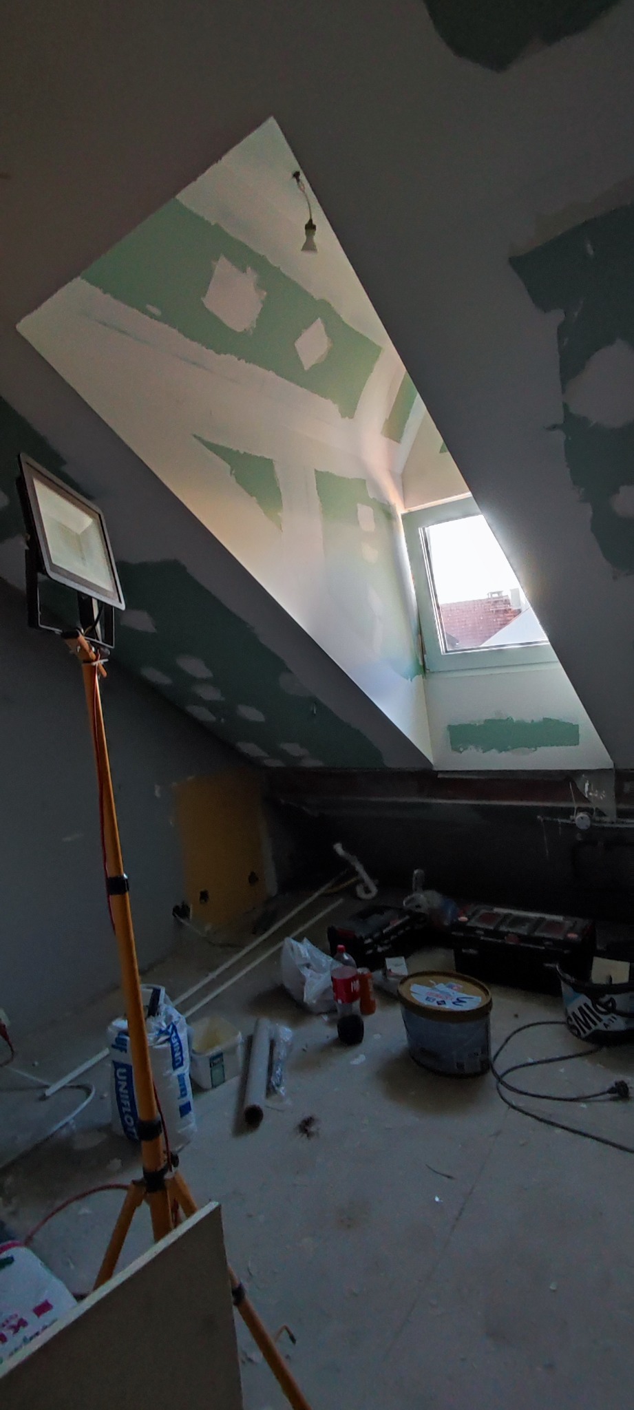 Poddasze w trakcie prac wykończeniowych: widoczne płyty kartonowo-gipsowe na skosach dachu, częściowo zaszpachlowane, reflektor budowlany na trójnogu oświetla wnętrze, na podłodze narzędzia...