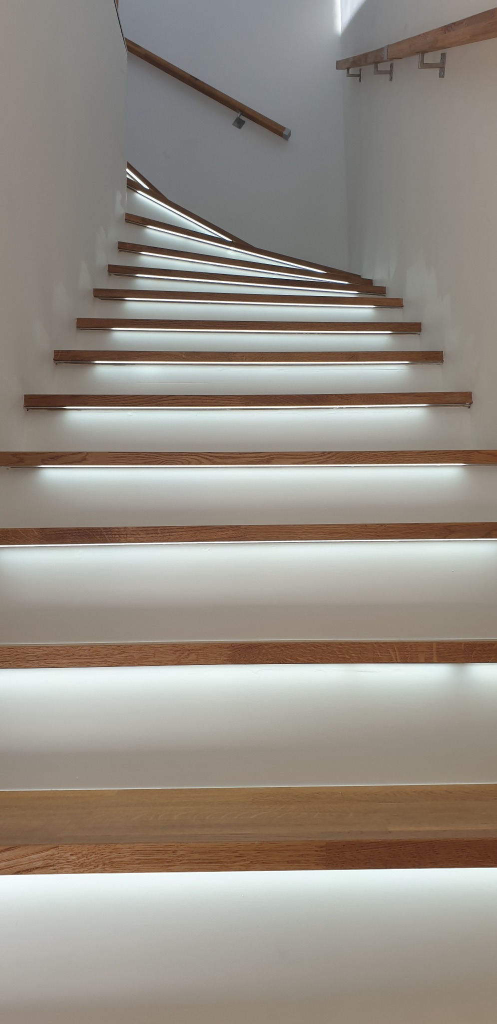 Drewniane schody ze zintegrowanym oświetleniem LED, prowadzące w górę, z minimalistyczną białą ścianą i drewnianą poręczą.