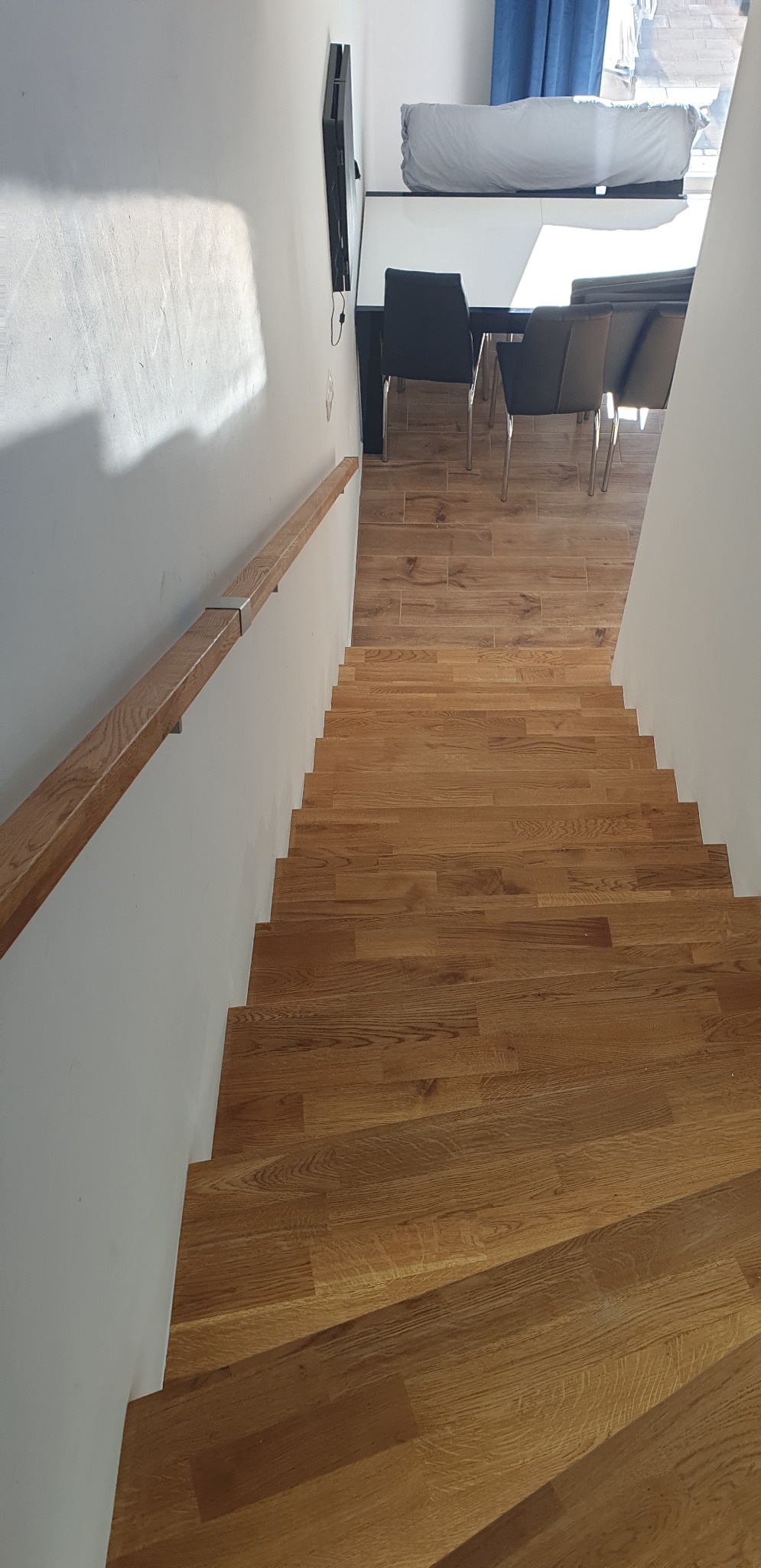 Schody z drewnianymi stopniami i białą balustradą, widok z góry, w tle fragment jadalni z krzesłami i stołem.