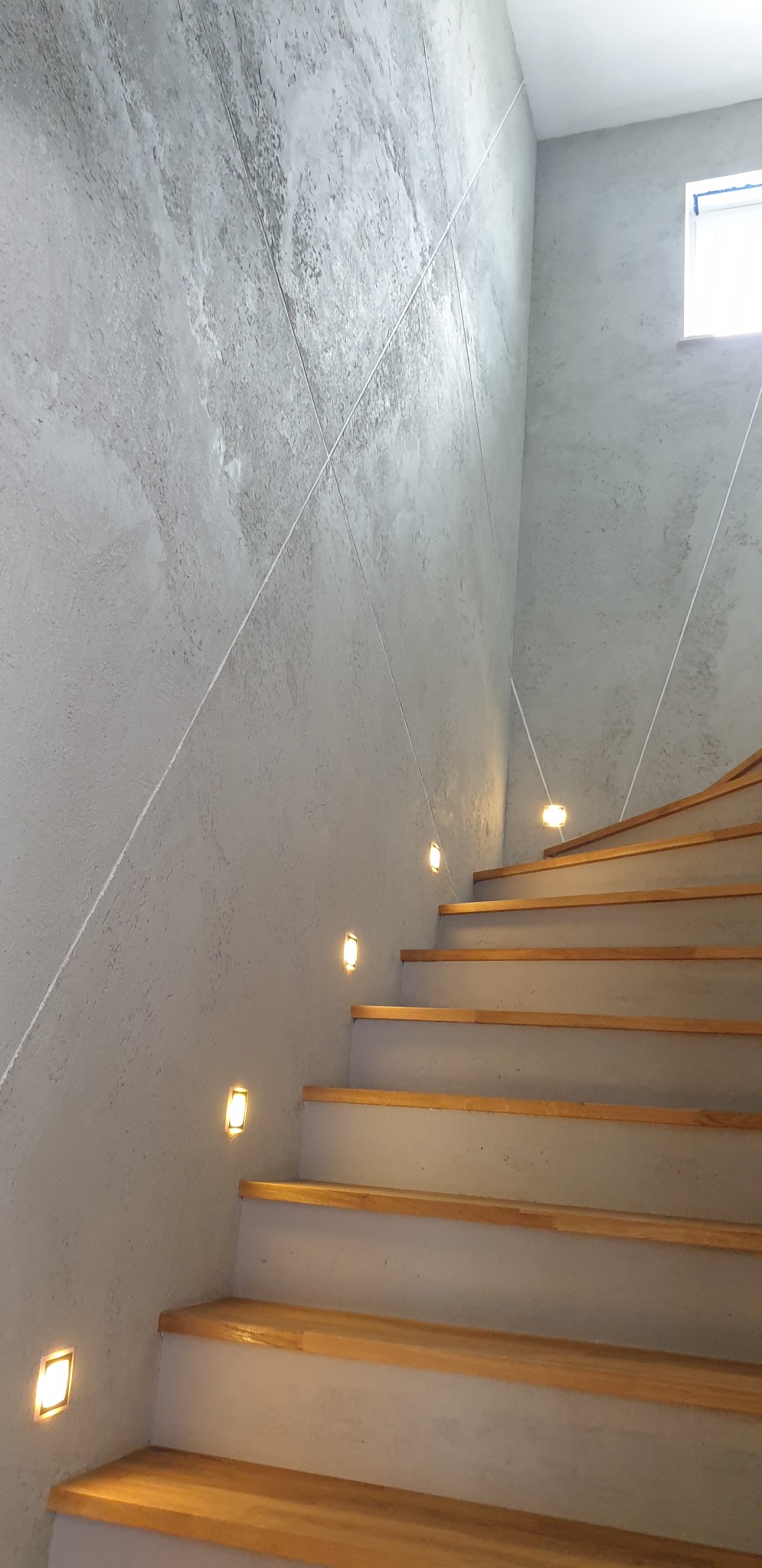 Schody drewniane z oświetleniem LED w ścianie o fakturze betonu architektonicznego, widok z dołu, minimalistyczny design.