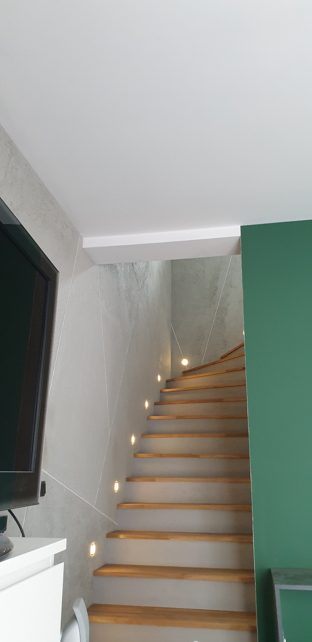 Klatka schodowa z drewnianymi stopniami i oświetleniem punktowym w ścianie o geometrycznym wzorze, widoczny fragment telewizora i zielonej ściany.