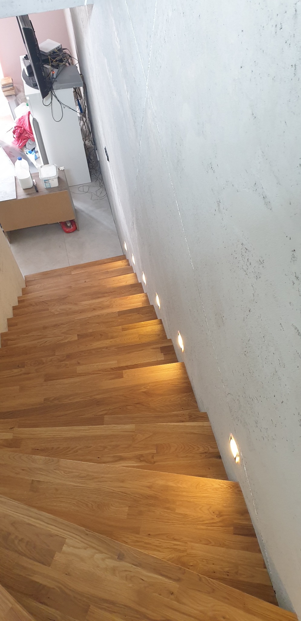 Drewniane schody z oświetleniem punktowym w ścianie o surowym wykończeniu, widok z góry, w tle niedokończone pomieszczenie z meblami i sprzętem RTV.