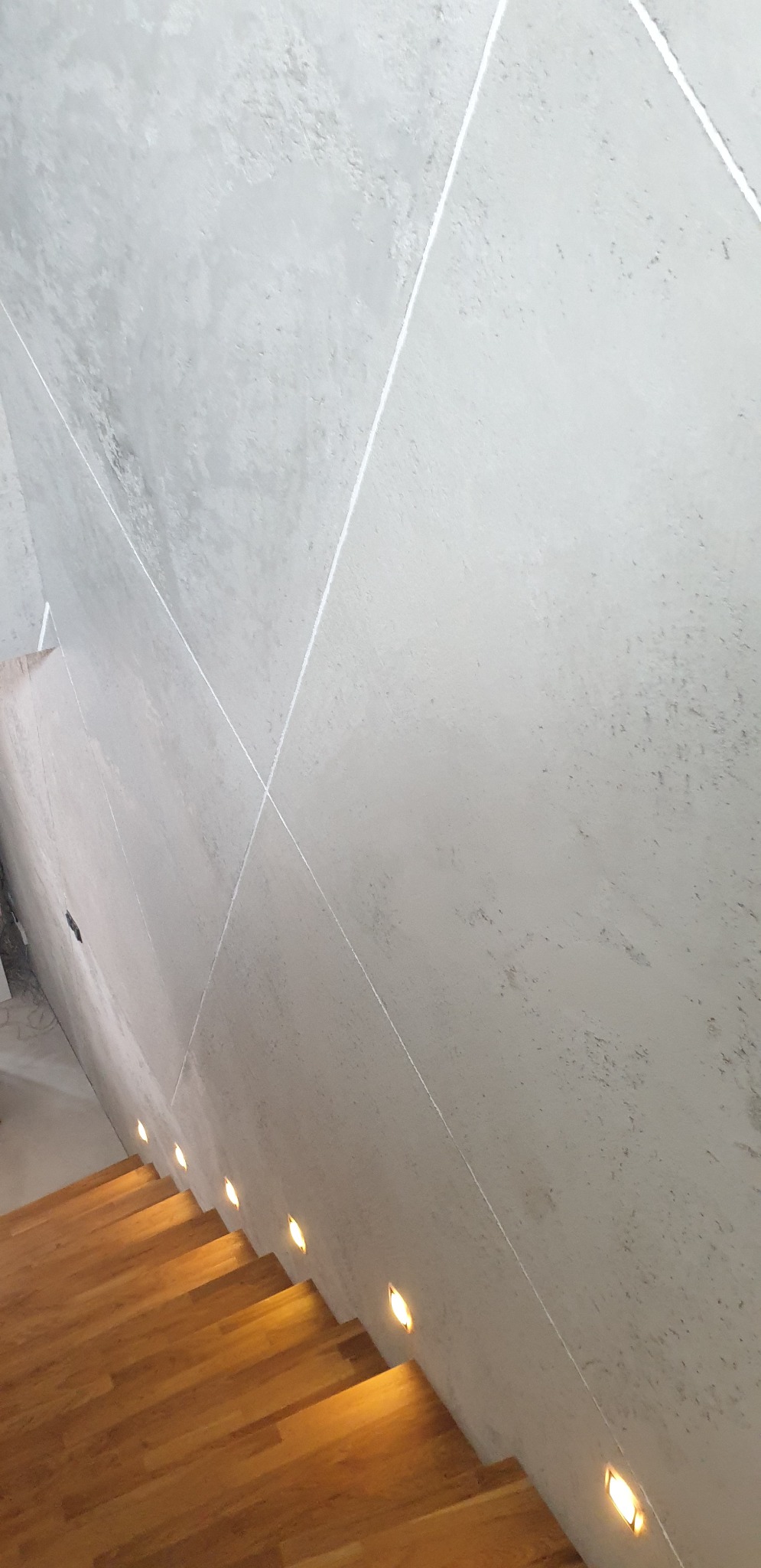 Betonowa ściana z krzyżującymi się liniami laserowymi nad drewnianymi schodami z wbudowanym oświetleniem punktowym.