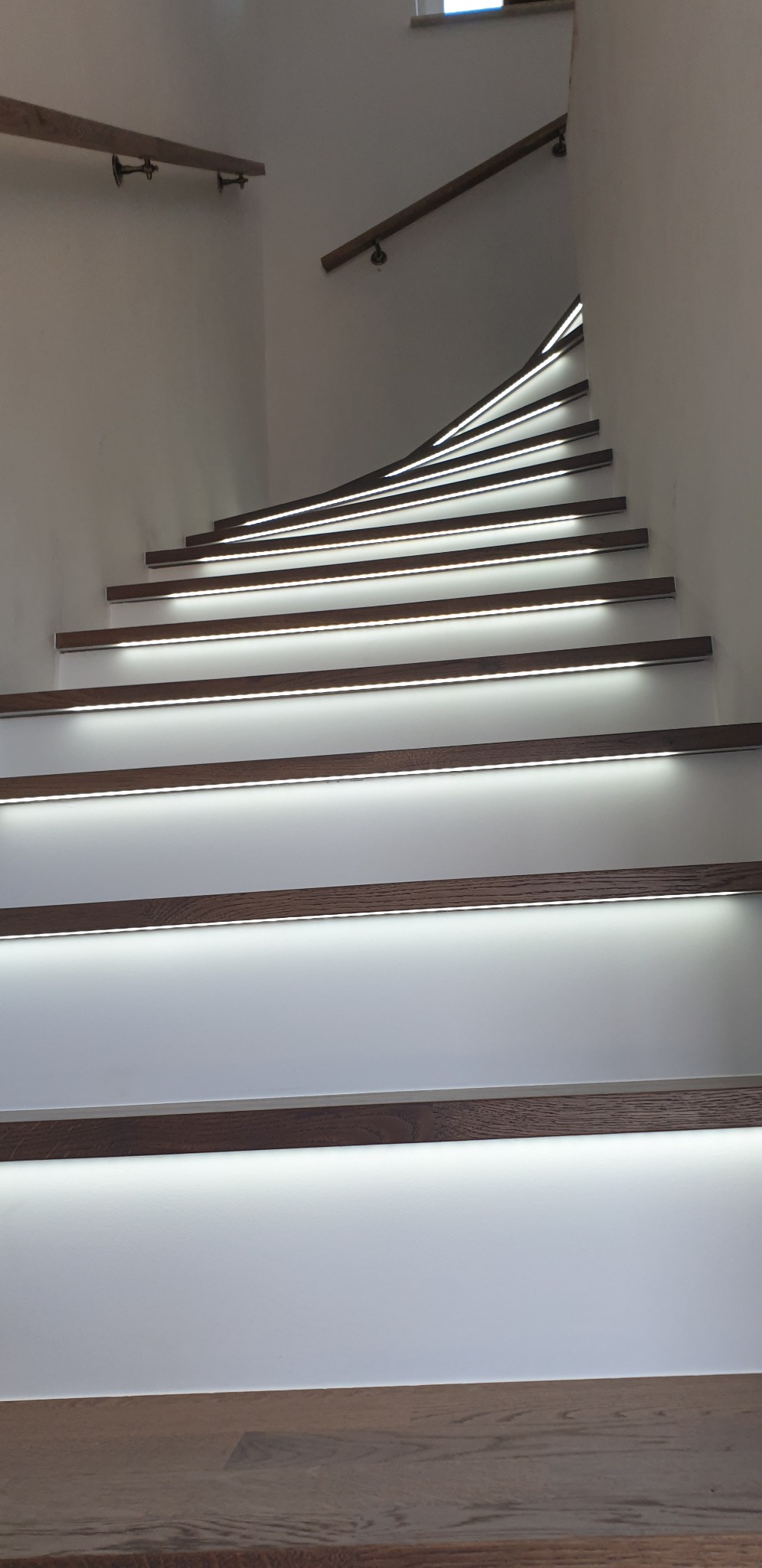 Nowoczesne schody z podświetleniem LED, drewniane stopnie i białe podstopnice, widoczne poręcze na ścianie.