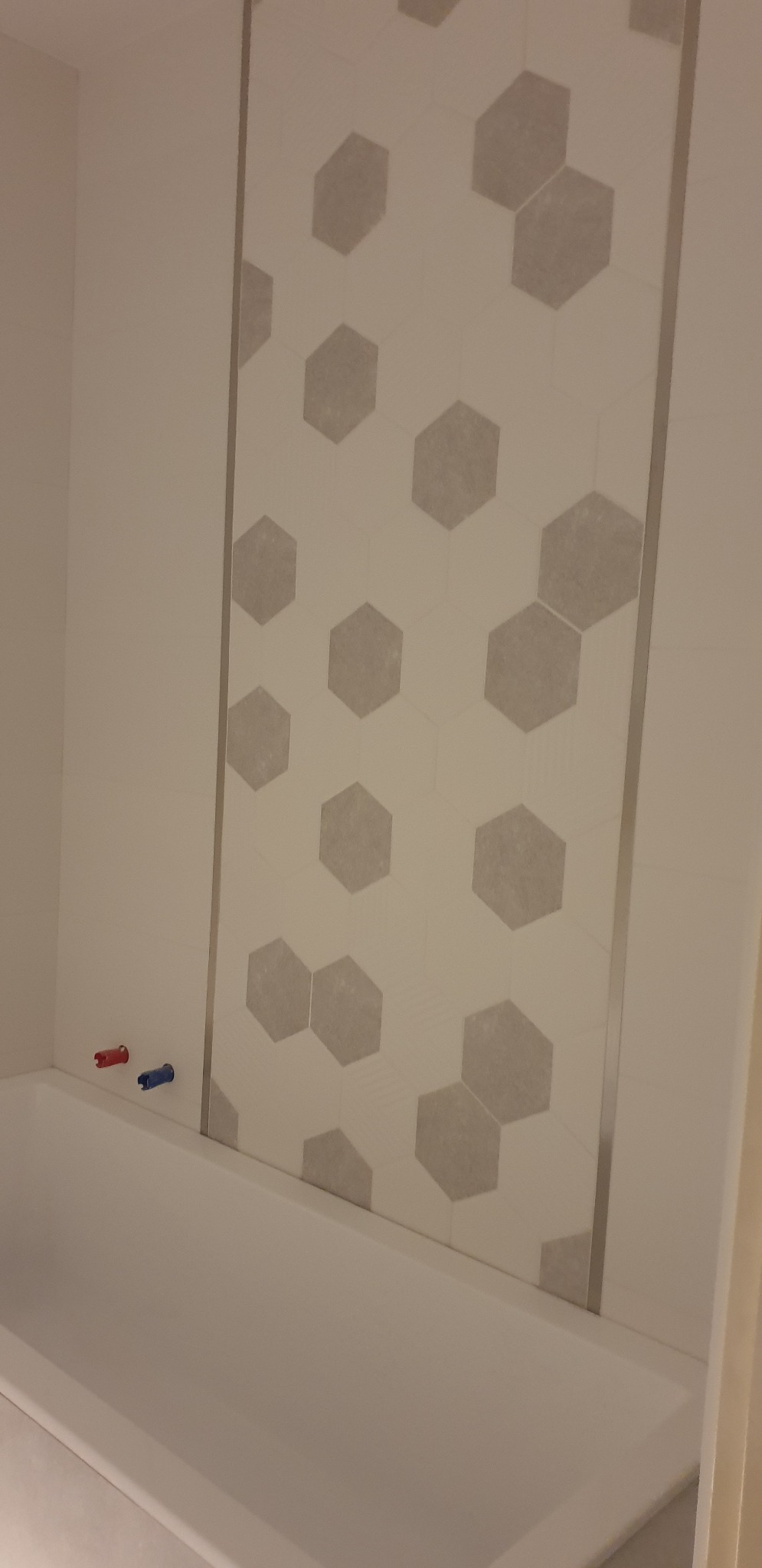 Fragment wykończonej łazienki: biała wanna, ściana wyłożona białymi heksagonalnymi płytkami z szarym wzorem, chromowana listwa wykończeniowa, wystające rury z zaworami w kolorze czerwonym...