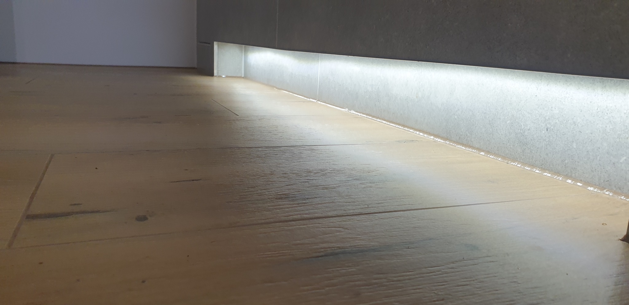 Zamontowane oświetlenie LED wzdłuż szarej ściany z płytek, nad drewnianą podłogą w pomieszczeniu w Swarzędzu.