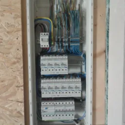 Rozdzielnica elektryczna natynkowa z zainstalowanymi bezpiecznikami i okablowaniem, widok z przodu na ścianie z płyt OSB i fragmentem ocieplenia.