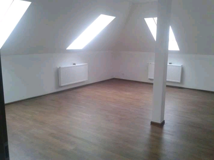 Poddasze po remoncie z drewnianą podłogą, białymi ścianami, grzejnikami i oknami dachowymi, wsparte na białym filarze.