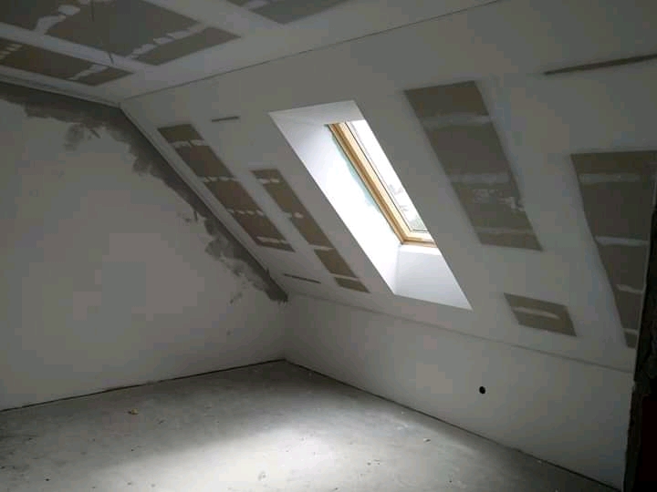 Poddasze w trakcie prac wykończeniowych, widoczne płyty kartonowo-gipsowe na skosach dachu i ścianie, okno dachowe w białej obudowie, oraz fragment ściany z naniesioną warstwą szpachli.