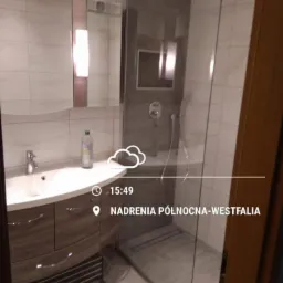 Nowoczesna łazienka po remoncie: kabina prysznicowa ze szklaną ścianką, umywalka z szafką w kolorze drewna i lustro z oświetleniem.