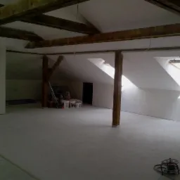 Poddasze po remoncie: białe ściany, drewniane belki stropowe, dwa okna dachowe wpuszczające jasne światło, widoczne narzędzia i materiały budowlane w tle.