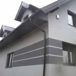 Dwukondygnacyjny dom z szarą elewacją i poziomymi pasami, widok z dołu na fragment ściany z oknami i rynnami.