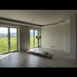 Wnętrze w trakcie prac wykończeniowych: szare ściany, jasne panele podłogowe, listwy sufitowe, duże okna z widokiem na zielone wzgórza, przenośne lampy budowlane oświetlające pomieszczenie, paczki...