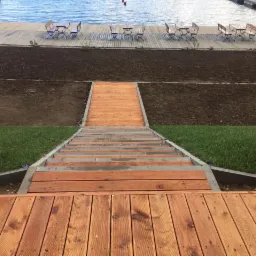 Drewniany taras z schodami prowadzącymi do pomostu nad wodą, z przygotowanym terenem pod trawnik i miejscami siedzącymi.