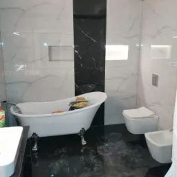 Elegancka łazienka z marmurowymi płytkami w czarno-białej kolorystyce, wolnostojąca wanna na ozdobnych nóżkach, podświetlane wnęki w ścianie, toaleta i bidet w minimalistycznym stylu.