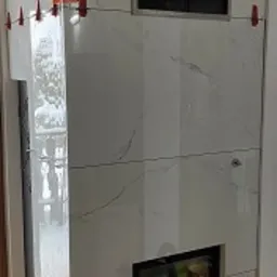 Montaż obudowy kominka z białych płyt imitujących marmur, widoczne pomarańczowe paski taśmy i czerwone klipsy poziomujące płytki.