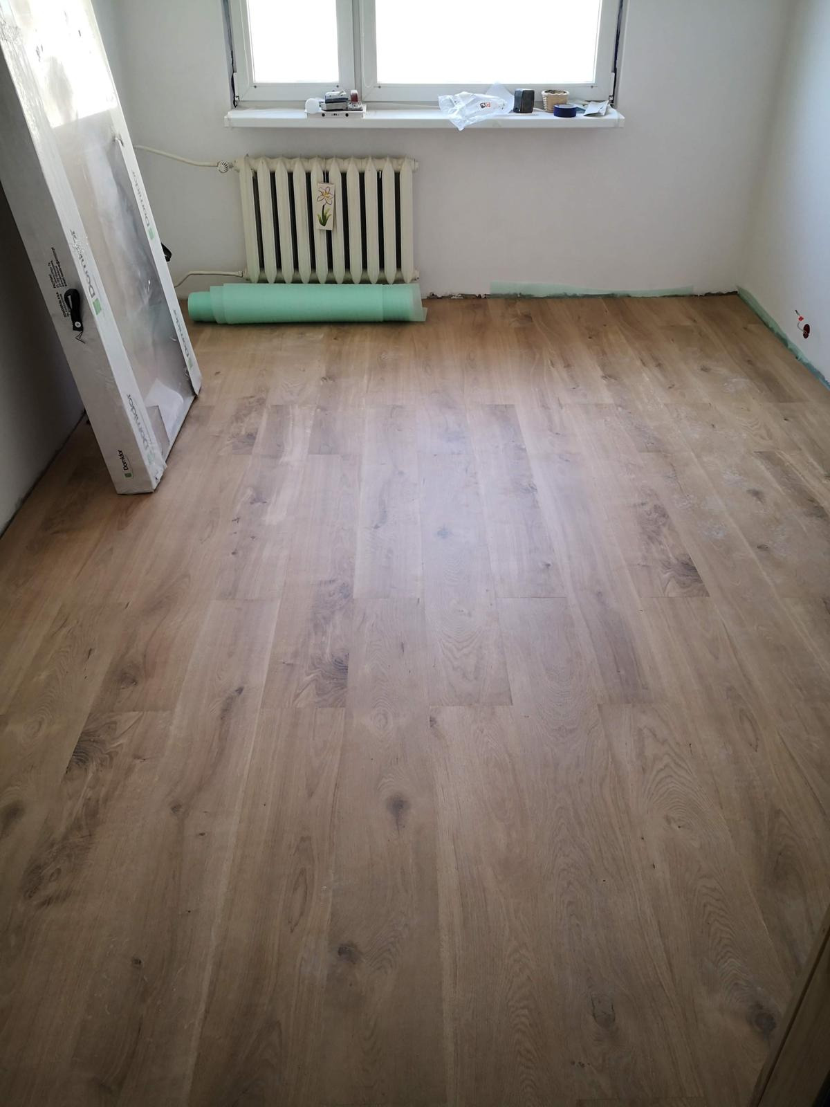 Układanie paneli podłogowych w pokoju, widoczny fragment nowej podłogi, rolka podkładu pod panele i zapakowane drzwi.