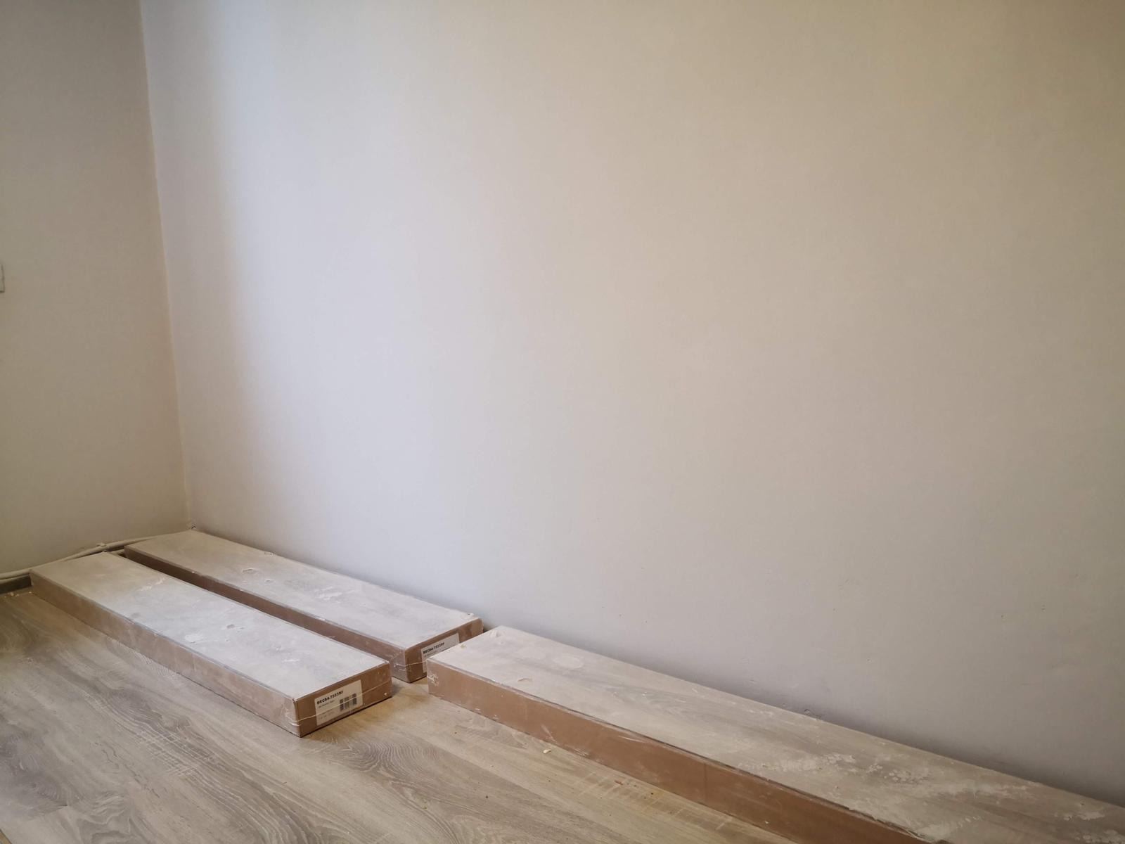 Paczki paneli podłogowych w kartonach, przygotowane do montażu przy białej ścianie na drewnianej podłodze.