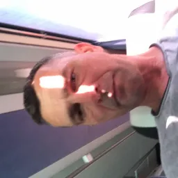 Portret mężczyzny w szarej koszulce, oświetlonego ostrym, jasnym światłem słonecznym padającym na twarz, z widocznym fragmentem wnętrza pomieszczenia w tle.