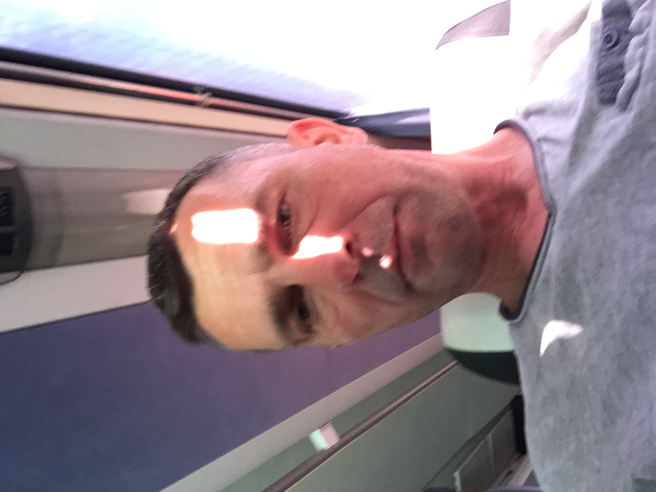 Portret mężczyzny w szarej koszulce, oświetlonego ostrym, jasnym światłem słonecznym padającym na twarz, z widocznym fragmentem wnętrza pomieszczenia w tle.