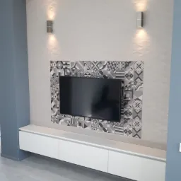 Szara ściana telewizyjna z dekoracyjnymi płytkami w geometryczne wzory, podświetlona dwoma kinkietami, z białą szafką RTV pod telewizorem.