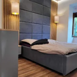 Sypialnia z łóżkiem z szarym tapicerowanym wezgłowiem z paneli, drewnianymi lamelami na ścianie i wiszącymi lampami z beżowymi abażurami. Widoczne okno z zielenią na zewnątrz.